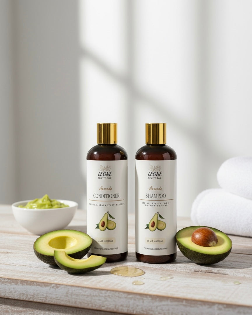 Two bottles of Leone Avocado hair care products on a wooden surface with avocados and a bowl of avocado butter.